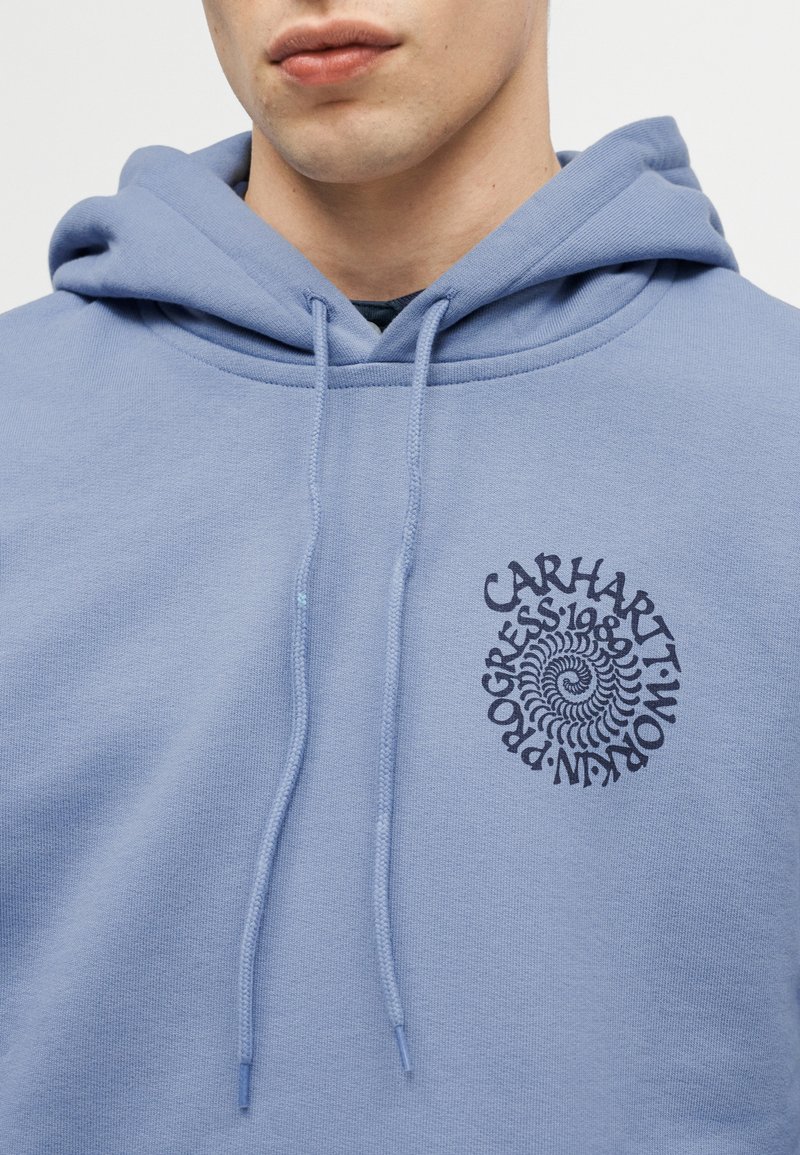 Persona joven con una sudadera con capucha Carhartt de color azul claro, con un logo de texto en espiral en el pecho y cordones colgando de la capucha.