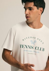 Biała bawełniana koszulka z napisem "Diverse Open Tennis Club" w zielonym i niebieskim kolorze, z grafiką skrzyżowanych rakiet. Krótkie rękawy i okrągły dekolt.