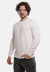 Maglione bianco lavorato a maglia con motivo testurizzato sul davanti, scollatura rotonda e polsini a costine, abbinato a jeans grigi scuri.