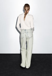 Světle modré džíny s volným střihem a viditelnými zadními kapsami, doplněné bílým topem s dlouhým rukávem, viděné zezadu.