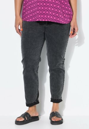 Femme portant un jean noir retroussé, des sandales slides noires et un haut rose à motifs, debout devant un fond blanc.