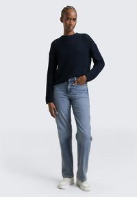 Vrouw met gevlochten haar, gekleed in een navy gebreide trui, lichtblauwe rechte jeans, witte sneakers, staand met één hand in de zak.