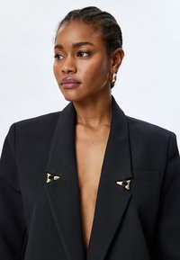 Zwarte blazer met een diepe V-hals, gestructureerde schouders en gouden hardware-accenten op de kraag. Soepele textuur en getailleerde pasvorm.