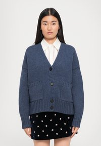 Sinine kudumine särk V-kaelusega, eesnuppe ja kaks ees taskut. Ribakudumisäärised ja -äär lisavad tekstuuri ja struktuuri.