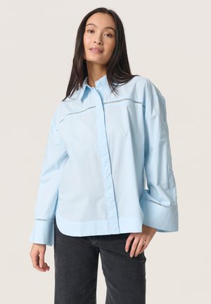 Light blue button-up shirt with a relaxed fit, featuring a stitched trim along the shoulders and bell-shaped sleeves.