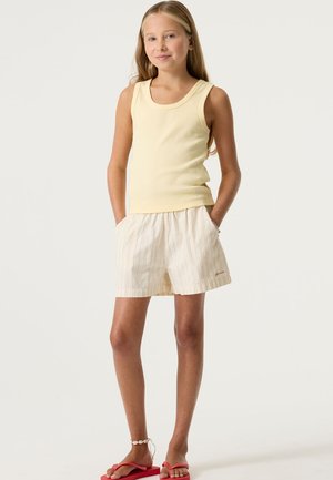 Geel mouwloos topje, gecombineerd met lichtbeige gestreepte korte broek met een elastische tailleband. Sandalen met rode bandjes, eenvoudig ontwerp.