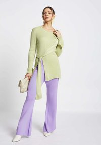 Maglione verde chiaro a coste con design avvolgente e fiocco in vita, abbinato a pantaloni a zampa d'elefante viola a coste e scarpe con tacco bianche. Borsa neutra.