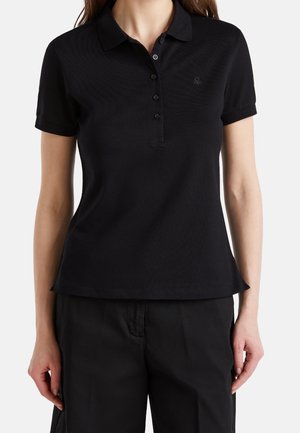 Frau trägt ein eng anliegendes schwarzes Poloshirt mit kurzen Ärmeln und Knöpfen sowie schwarze Hose, steht vor einem schlichten Hintergrund.
