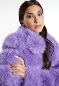 Lila Kunstfellmantel mit weicher, flauschiger Textur; verfügt über einen übergroßen Kragen und abgerundete Details. Model trägt Ohrringe, die Hände sanft übereinandergelegt.