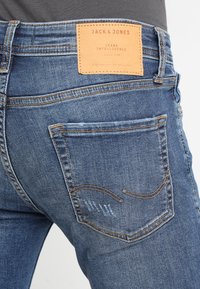 Blå denimjeans med en brun läderetikett vid midjan, med en klassisk fickdesign, sömdetaljer och en urtvättad look.