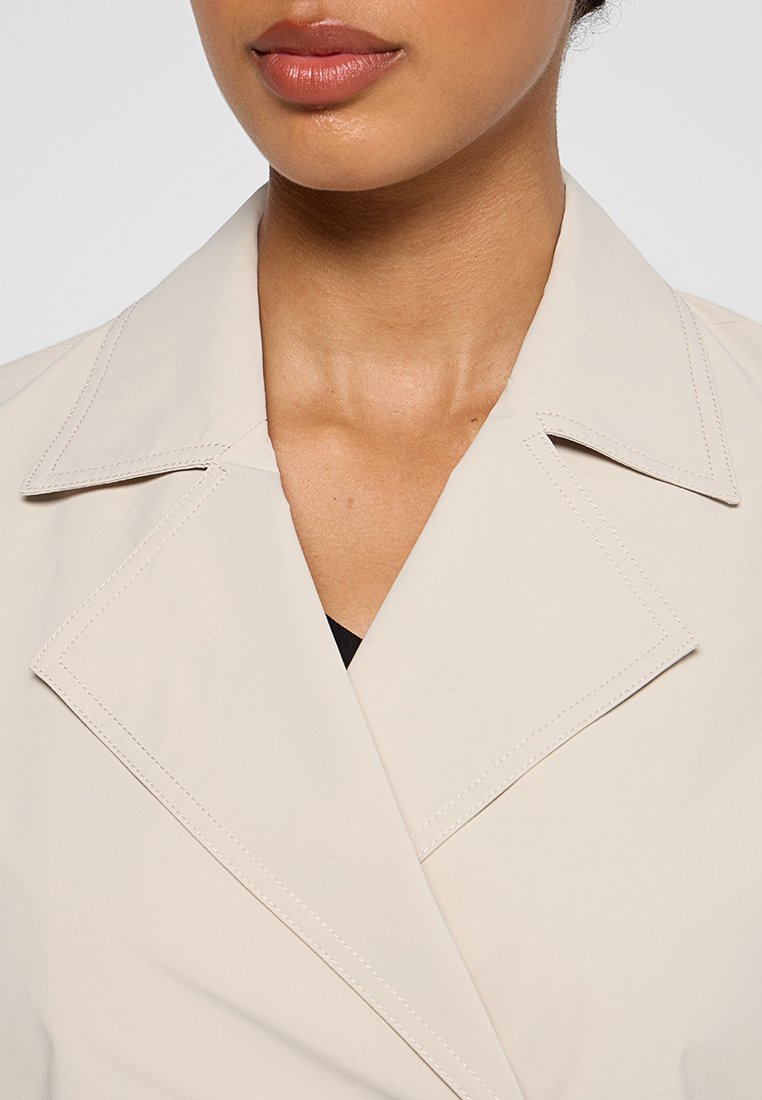 Trench beige con un amplio cuello de muesca y detalles de costura visibles. La tela parece suave y ligera, diseñada para superponerse.