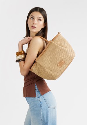 Donna che indossa un top marrone senza maniche e jeans azzurri, porta una grande borsa tote beige intrecciata sulla spalla, di profilo.