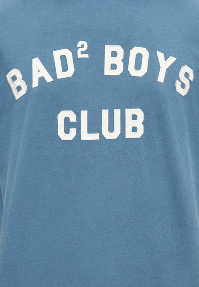 Μπλε ύφασμα με ανάγλυφα λευκά γράμματα που σχηματίζουν τη φράση «BAD² BOYS CLUB» σε καμπυλωτή και ευθεία διάταξη.