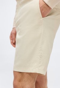 Μπεζ σορτς μέχρι το γόνατο σε άτομο με το ένα χέρι στην πλαϊνή τσέπη, με το λογότυπο "Oakley" κεντημένο κοντά στο τελείωμα.