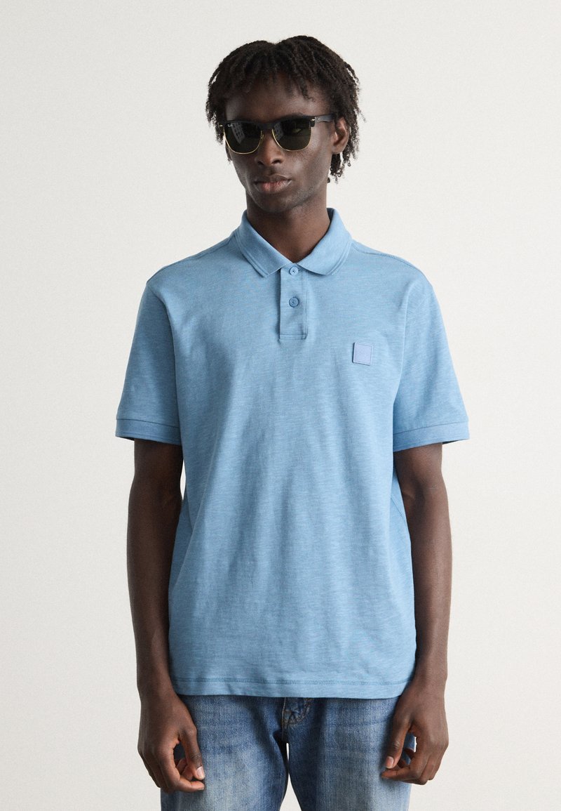 Polo azul claro com acabamento texturizado, com pala de botão de três botões e um pequeno emblema. Modelo a usar óculos escuros escuros e jeans.
