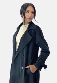 Abrigo negro de trench con grandes solapas, diseño de doble botonadura, bolsillos laterales y puños ajustables. Lleva puesto un suéter de canalé de color beige claro.