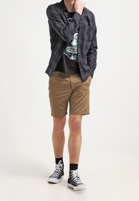 Conjunto informal que incluye una camisa de cuadros negra, una camiseta gráfica, pantalones cortos marrones y zapatillas de lona negras con suelas blancas y calcetines negros.