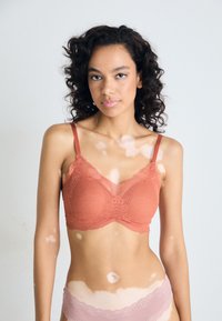 Blonde bralette i korall med justerbare stropper, har et semi-transparent design og bølgende kanter, kombinert med lyse rosa undertøy.