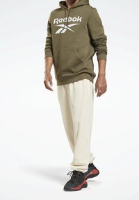 Olivgrüner Hoodie mit weißem Reebok-Logo, kombiniert mit locker sitzenden, cremefarbenen Jogginghosen und schwarzen Turnschuhen mit roten Akzenten.