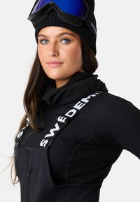 Sort skioutfit med ribbet langærmet trøje, fleece halsvarmer og stropdetaljer. Accessories med sort beanie og skigoggles.