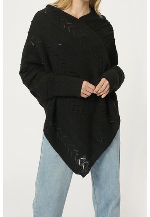 Frau trägt einen schwarzen Strickponcho mit Chevron-Mustern und langen Ärmeln, kombiniert mit hellblauen Jeans.
