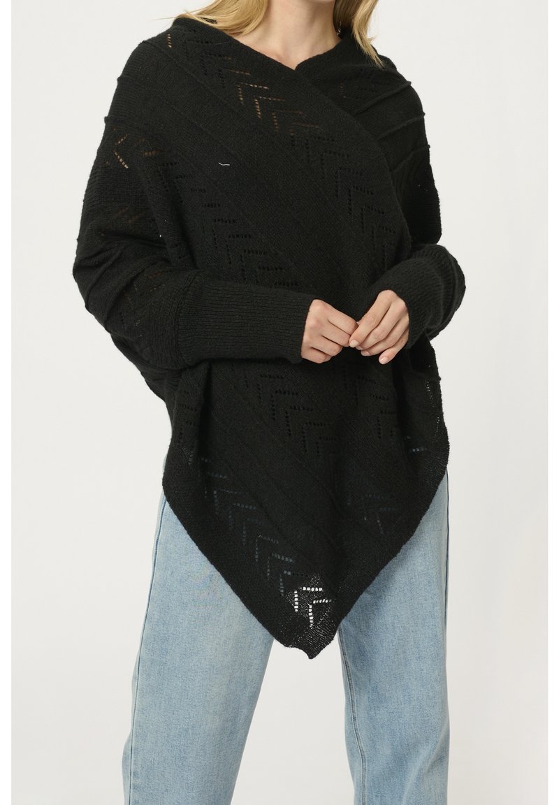 Femme portant un poncho en maille noire à motifs chevrons et manches longues, associé à un jean en denim bleu clair.