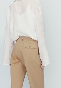 Chemise en lin blanc légère et transparente avec des manches longues, associée à un pantalon en coton beige clair avec une poche arrière et des détails de boutons.