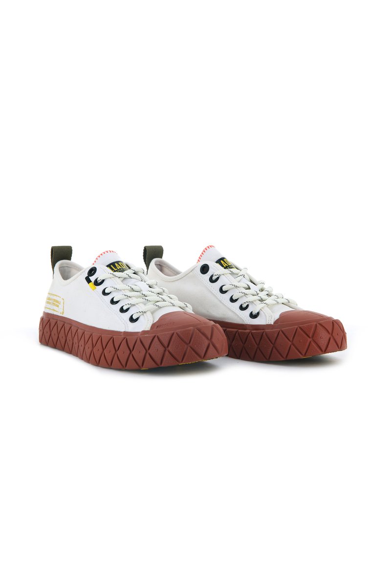 Baskets en toile blanches avec une semelle en caoutchouc marron présentant un motif en diamant texturé, des œillets noirs et des accents contrastants jaunes et rouges.
