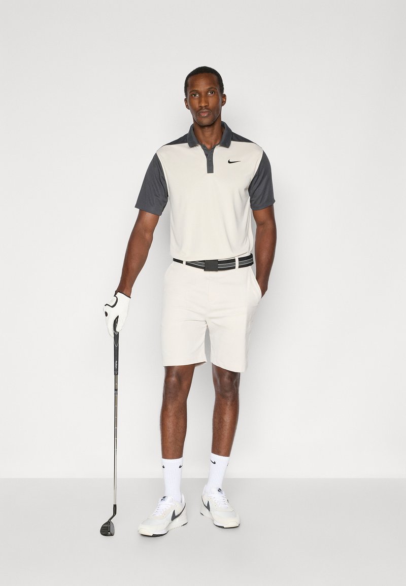 Weißes und graues Golfpolo mit Kragen und Knopfleiste. Helle graue Shorts und weiße Schuhe. Hält einen Golfschläger und trägt einen weißen Handschuh.
