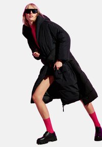 Cappotto puffo nero con cappuccio oversize, chiusura a zip e vestibilità ampia. Abbinato a un maglione rosa, calze rosa e scarpe nere chunky.