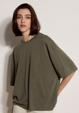 Jonge vrouw met kort bruin haar draagt een oversized olijfgroene t-shirt, staand tegen een effen beige muur met een neutrale uitdrukking.