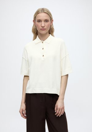 Γυναίκα με ξανθά μαλλιά που φοράει κρεμ polo μπλούζα με κοντά μανίκια και σκούρα καφέ παντελόνια, στέκεται μπροστά σε ένα απλό λευκό φόντο.