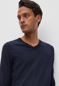 Tamsiai mėlynas V formos megztinis, pagamintas iš minkštos mezgimo medžiagos. Pristatomi suplakti akcentai prie kaklo ir rankogalių, su siauru, prigludusiu siluetu.