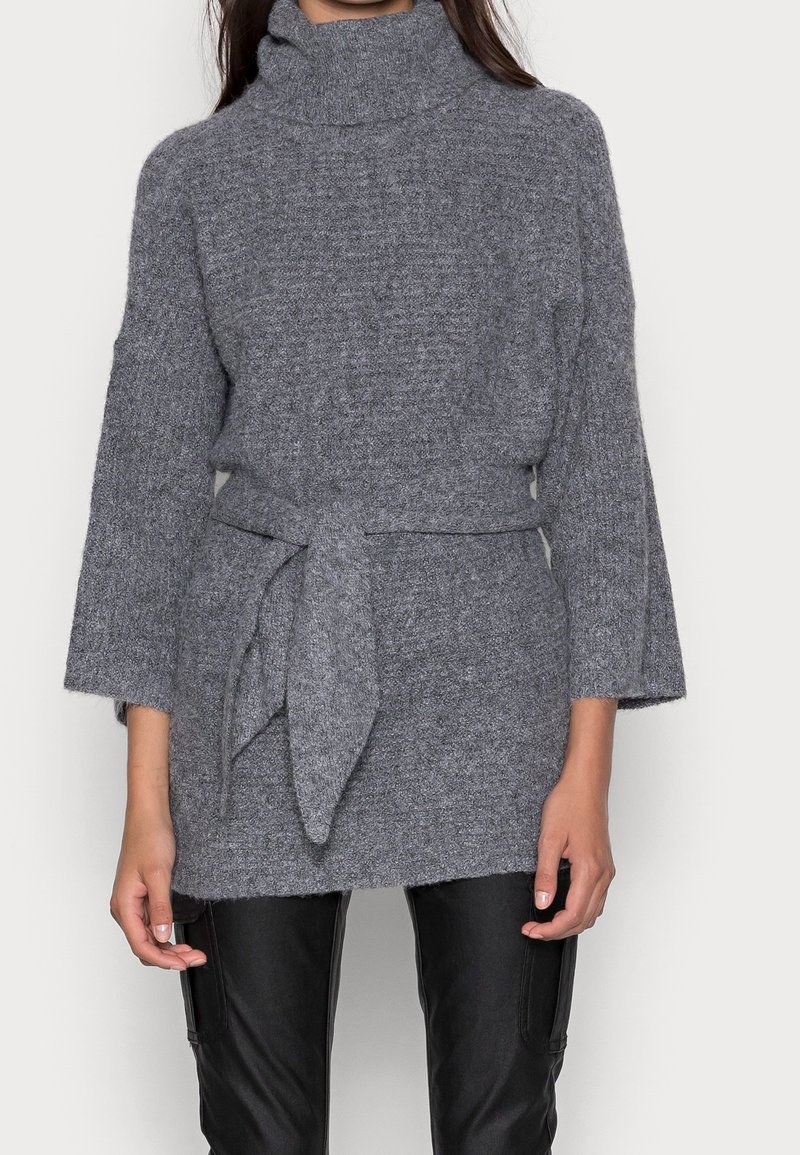 Frau trägt einen grauen gestrickten Rollkragenpullover mit einem breiten Gürtel, der um die Taille gebunden ist, und schwarze Lederhosen, stehend vor einem weißen Hintergrund.