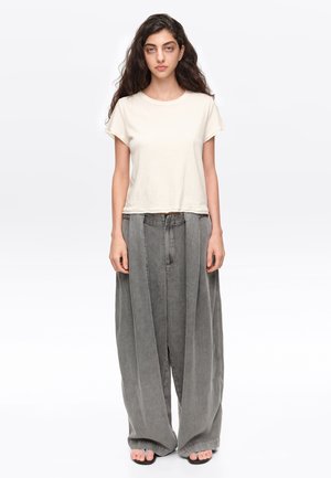 Mujer con cabello largo y oscuro que lleva una camisa blanca de manga corta y pantalones anchos grises extragrandes, de pie descalza sobre un fondo blanco.