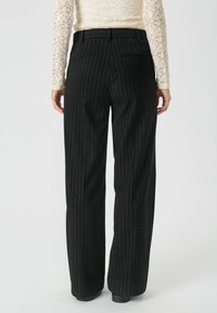 Pantalons noirs à rayures avec une coupe large, présentant une taille haute et deux poches au dos. Le tissu semble lisse avec un éclat subtil.