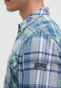 Karobluse in Blau-, Grün- und Grautönen. Mit Kragen, Knopf-Taschen und einem Marken-Patch auf dem Ärmel. Strukturierter Stoff.