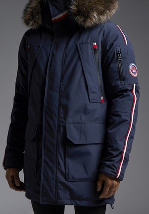 Parka marine avec une capuche en fausse fourrure, dotée de multiples poches zippées et d'accents rayés rouges et blancs le long des manches.