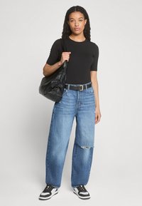 Black fitted top, faded blue wide-leg jeans with a tear, black belt with gold buckle, carrying a black slouchy handbag, and white sneakers.