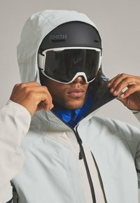 Ski jacket in light blue with a black zipper, worn over a blue top. Features a hood and a black helmet with goggles, showcasing sleek design.