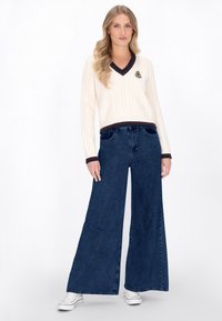 Cremfarbener Kabelstrickpullover mit tiefem V-Ausschnitt und dunkler Kante, kombiniert mit weiten, dunkelblauen Jeans und weißen Sneakern.