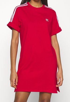 Kobieta ubrana w czerwony T-shirt-sukienkę Adidas z białymi paskami na ramionach i logo na piersi, stojąca z rękami swobodnie opuszczonymi wzdłuż ciała.