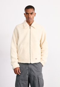 Giacca in pile color crema con zip anteriore, colletto e tasche laterali; presenta una superficie texturizzata e ricamo del marchio sull'orlo.