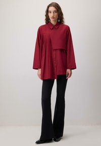 Bordo värvi pluus nööpidega kaeluse, vertikaalsete voldikute ja kihilise kangadisainiga, mida on kombineeritud mustade, kitsaste ja lehvivate pükstega ning mustade jalatsitega.