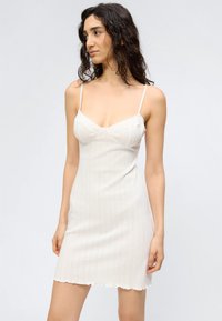 Γυναίκα με κυματιστά σκούρα μαλλιά, ντυμένη με άσπρο, ριπ πλεκτό φόρεμα χωρίς μανίκια με λεπτές τιράντες, στέκεται μπροστά σε απλό φωτεινό φόντο.