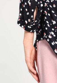 Schwarze Bluse mit weißen und korallenfarbenen Punkten, mit umgeschlagenem Ärmel und Cut-out-Detail. Weicher, leichter Stoff. Rosa Hose teilweise sichtbar.