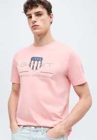 Νεαρός με κοντά, σγουρά μαλλιά, που φοράει ροζ μπλουζάκι GANT με λογότυπο ασπίδας και ανοιχτόχρωμο τζιν, με τα χέρια στις τσέπες, σε ουδέτερο φόντο.