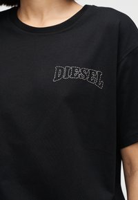 Άτομο που φοράει μαύρο t-shirt με στρογγυλή λαιμόκοψη και λευκό κεντημένο λογότυπο "DIESEL" στο πάνω αριστερό μέρος του στήθους.