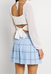 Haut blanc à manches longues avec découpe au dos et fermeture à nouer, associé à une jupe plissée bleu clair avec un motif quadrillé en lignes noires.