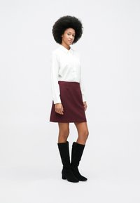 Žena stoji s kovrčavom kosom, odjevena u bijelu bluzu dugih rukava, burgundan suknju i crne čizme do koljena na praznoj pozadini.